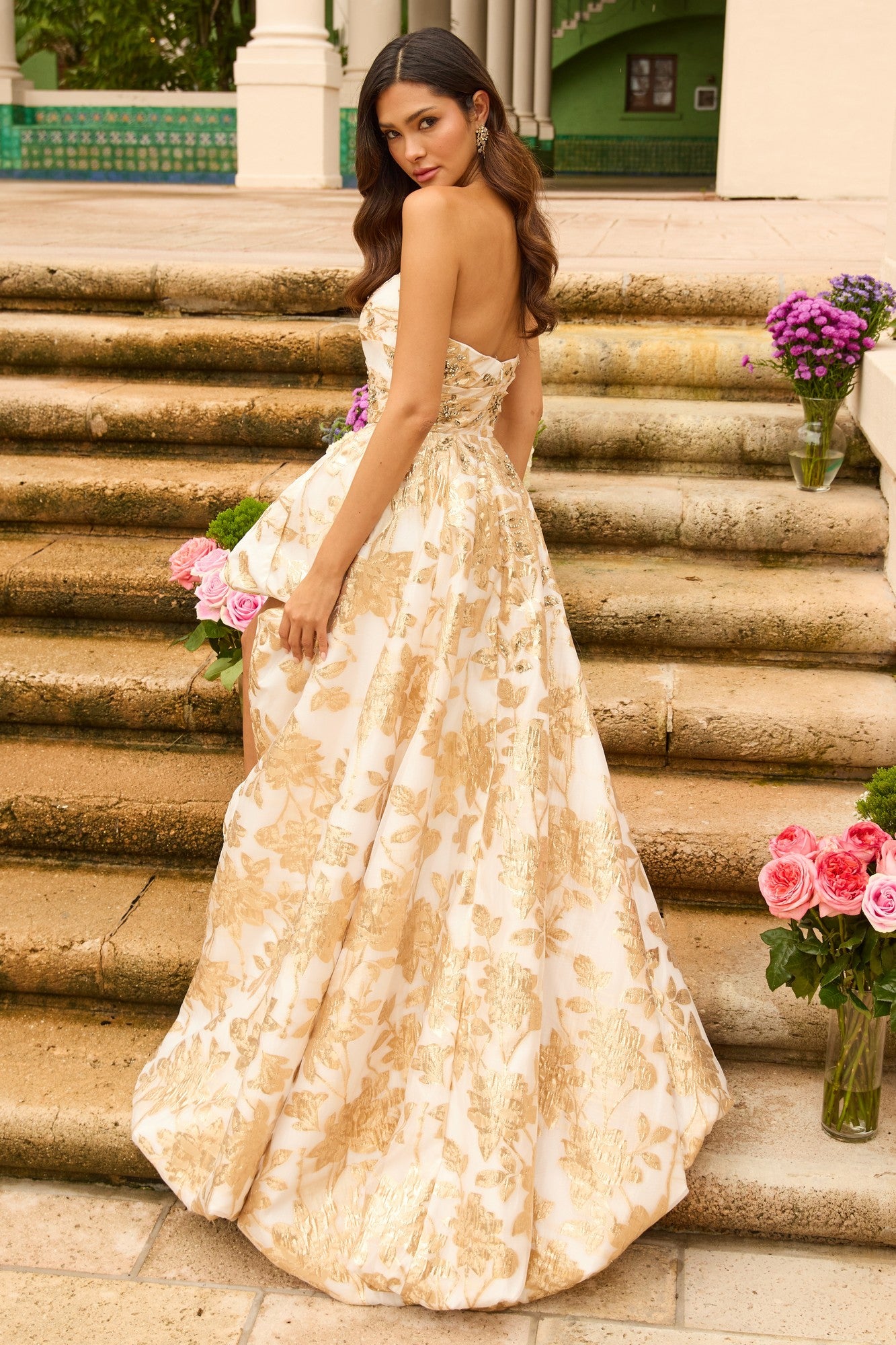 Wearing the Ava Presley 42344 long prom dress, a woman stands on stone steps holding flowers, with pink roses in vases beside her.